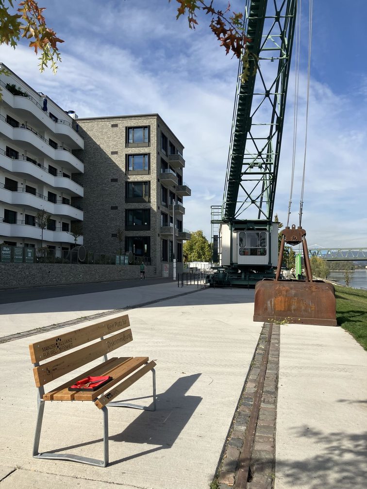 Parkbank gegen Diskriminierung der Allianz für Weltoffenheit und der Landeshauptstadt Mainz am Ufer des Zollhafens. Im Hintergrund ein alter Kran sowie Wohnhäuser.