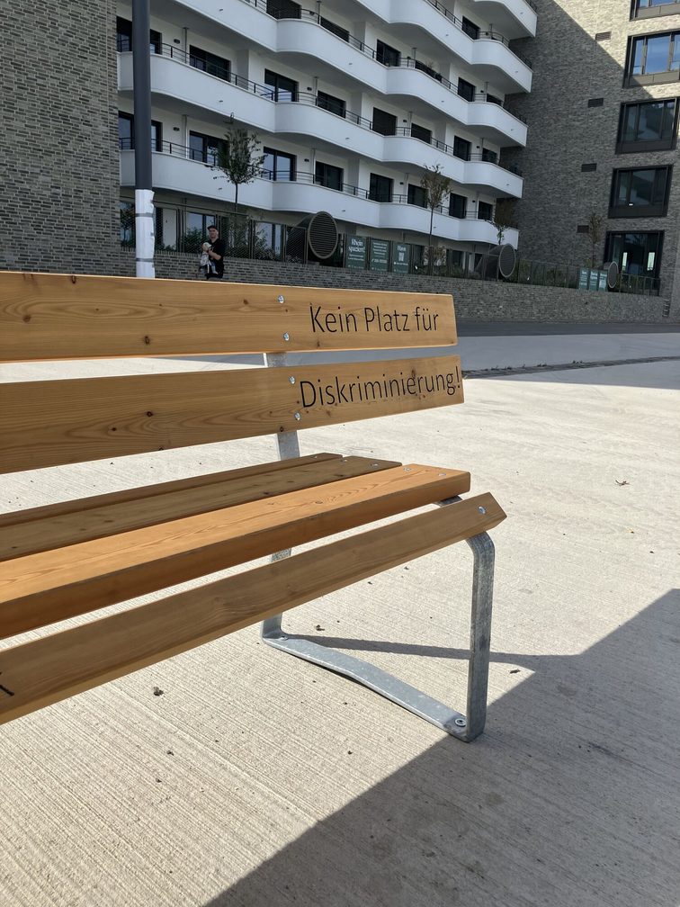 Bank mit Aufschrift "Kein Platz für Diskriminierung" auf der Banklehne. Davor fehlt an dieser Stelle die Sitzfläche