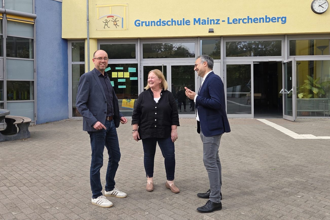 Christian Hoffmann, kierownik działu ds. klientów i relacji z opinią publiczną w Mainzer Mobilität, Susanne Millen, dyrektorka szkoły podstawowej Mainz-Lerchenberg oraz Ata Delbasteh, dyrektor ds. edukacji w stolicy kraju związkowego Moguncji, stoją przed budynkiem szkoły z napisem „Szkoła podstawowa Mainz-Lerchenberg”