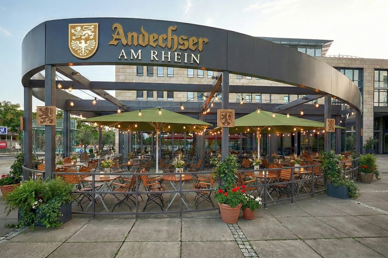 الجزء الخارجي لمطعم «Andechser am Rhein»: توجد العديد من الكراسي تحت مظلات خضراء، وتزين سلاسل الأضواء هذه المنطقة.