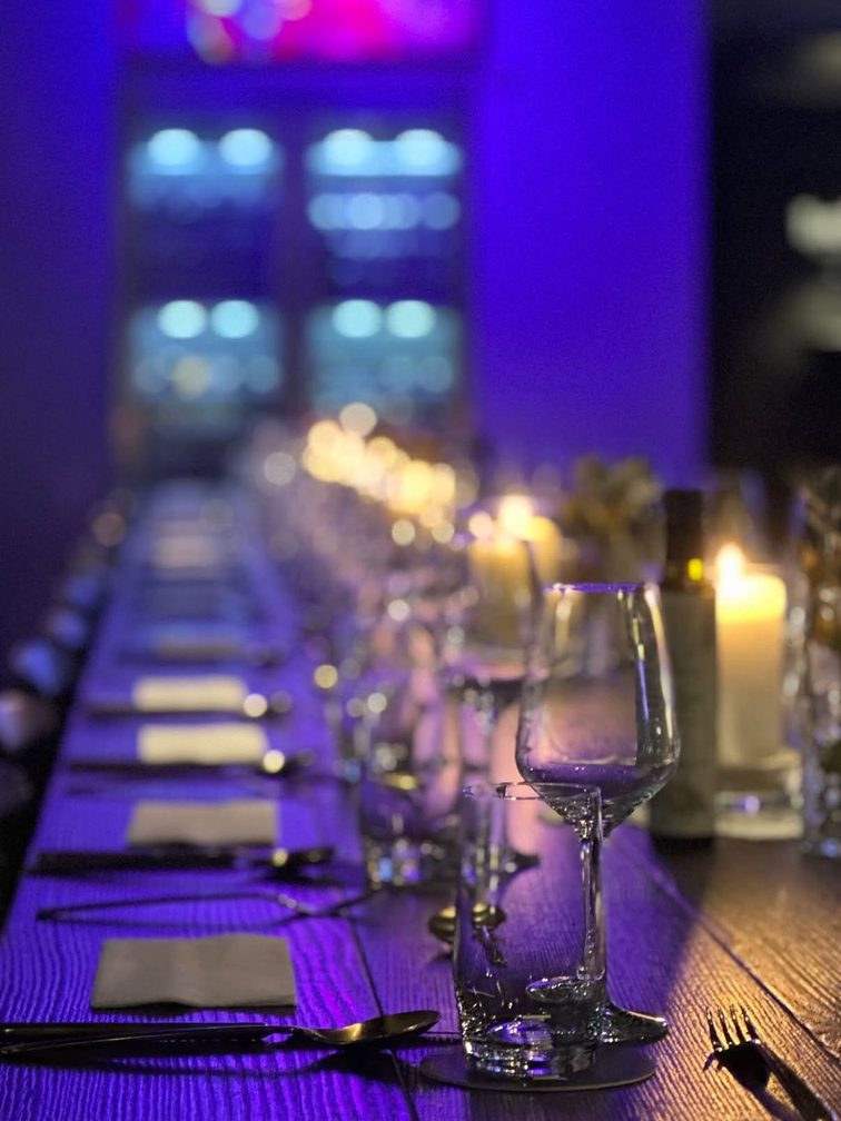 Au premier plan, on aperçoit une table dressée qui invite à s'attabler. Le mur est éclairé d'une lumière bleue et de nombreuses bougies sont allumées sur la table.