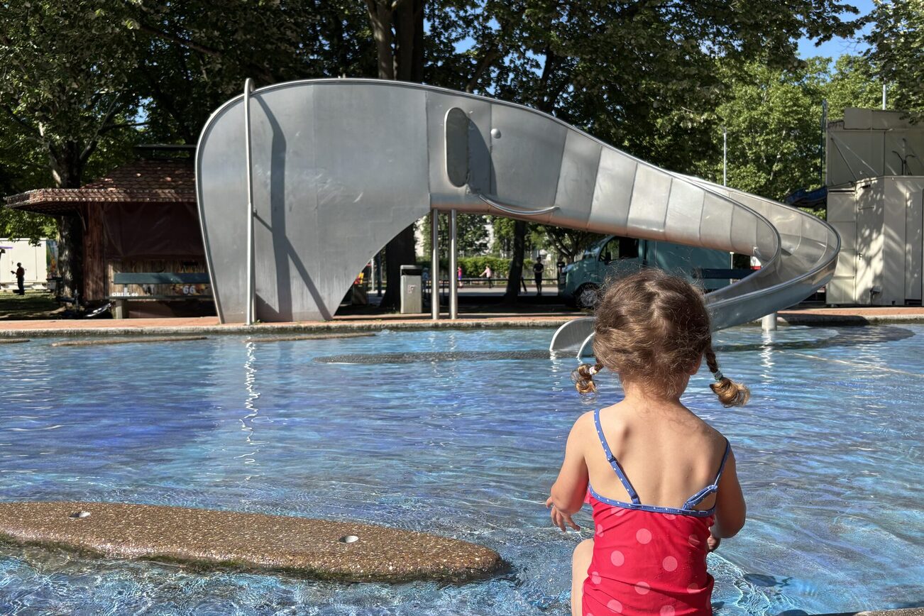 Ein Mädchen in einem rot gepunkteten Badeanzug sitzt am Wasserbecken und schaut zu einer Rutsche, die die Form eines Elefanten hat.