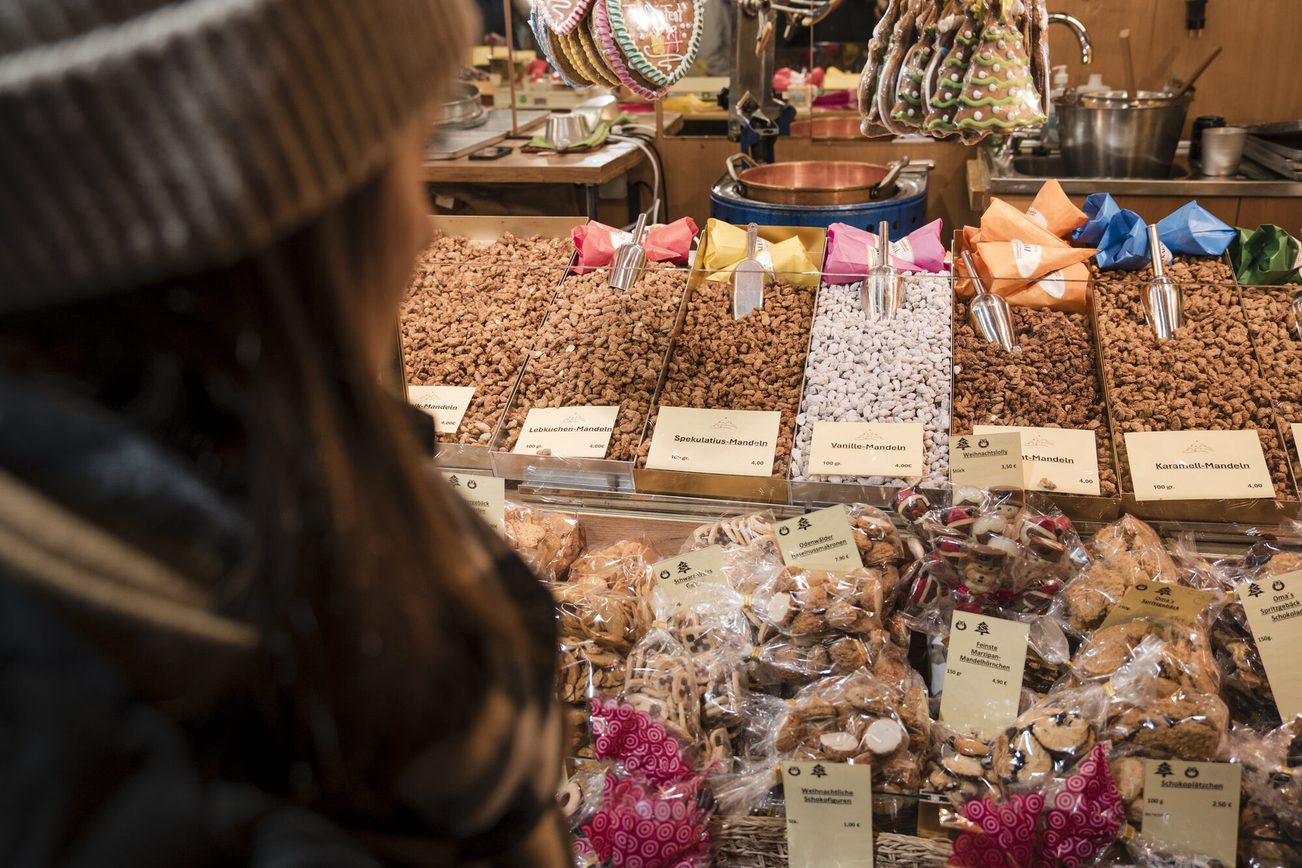 Rückenansicht einer Person mit Mütze, Schal und Jacke, die sich einen Süßigkeitenstand auf dem Weihnachtsmarkt anschaut