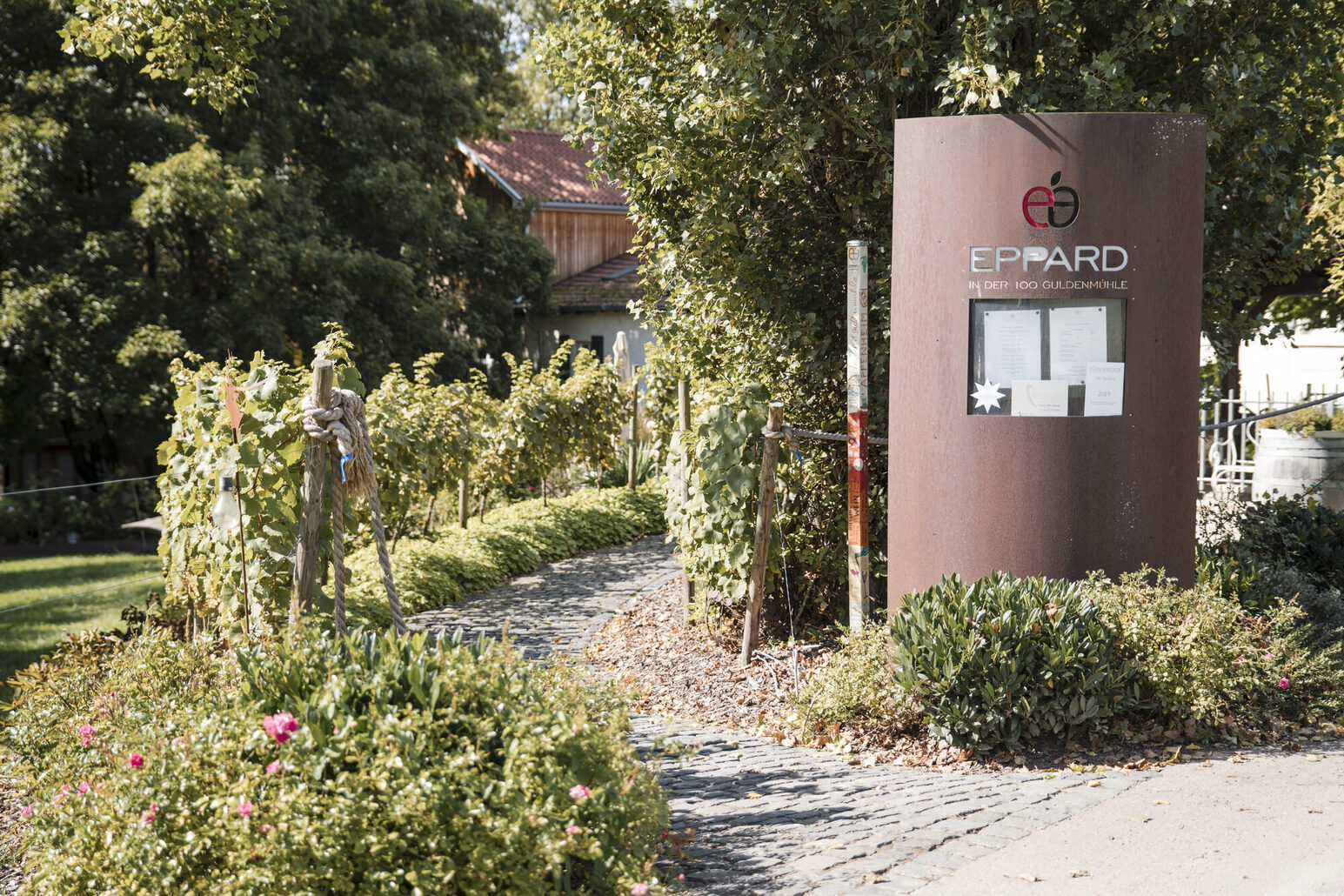 Ein Bild zeigt den begrünten Weg zum Eingang des Restaurants, rechts eine Stele mit der Aufschrift Eppard und Aushängen.