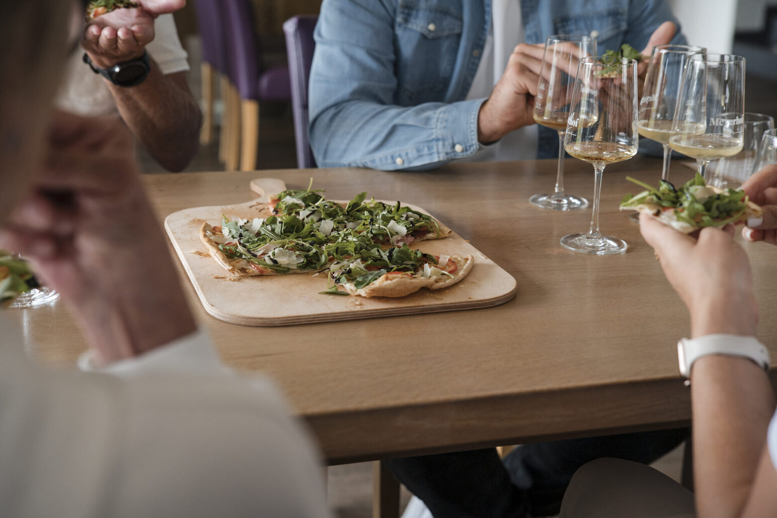 Ein gedeckter Tisch mit Flammkuchen und Hände, die Flammkuchenstücke halten. Auf dem Tisch stehen vier Gläser mit Weißwein.