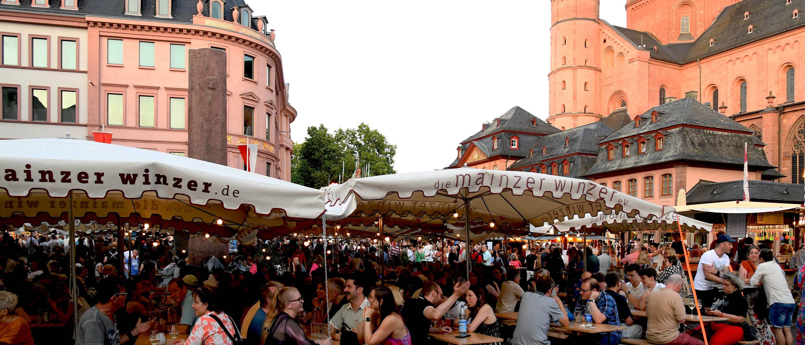 Menschen sitzen auf dem Markt auf Bierbänken unter Sonnenschirmen der Mainzer Winzer auf dem Fest der Mainzer Johannisnacht. Rechts im Hintergrund ist der Dom zu sehen.