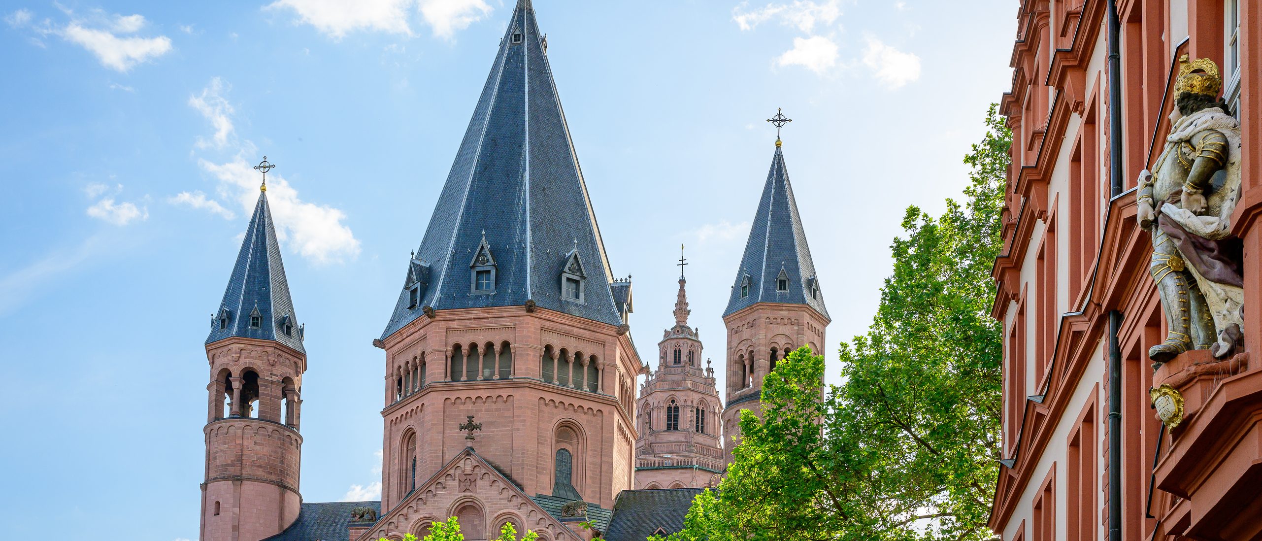 Im Mittelpunkt der Ostchor des Doms in rotem Sandstein, davor grüne Baumkronen, rechts das Haus zum Römischen Kaiser, weiß mit roten Elementen