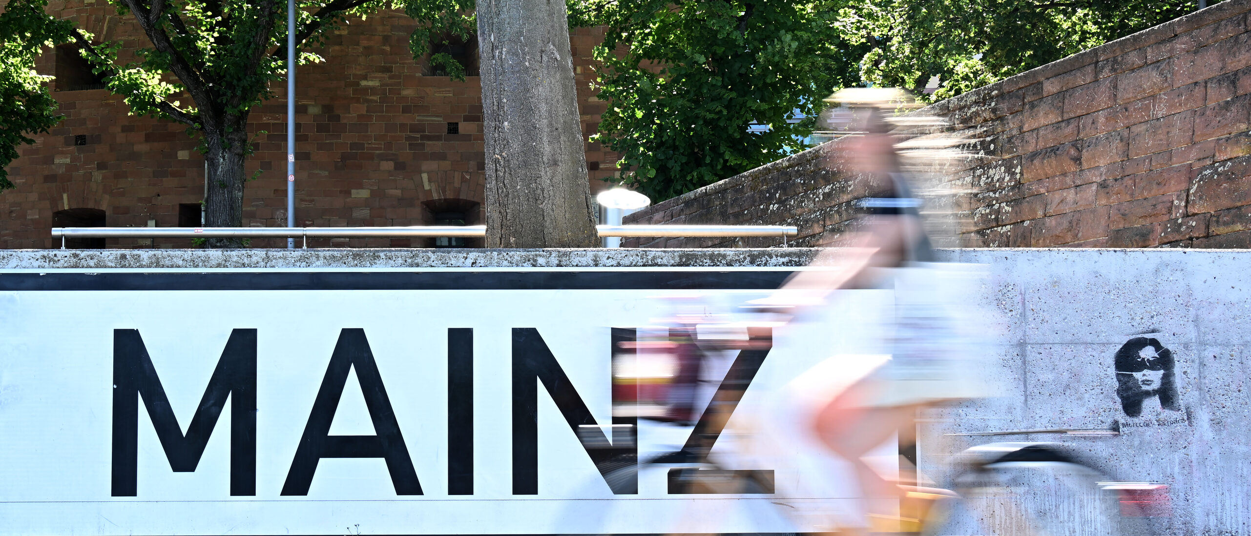 Schriftzug MAINZ in Großbuchstaben, schwarz auf weiß auf grauer Mauer, davor eine Radfahrerin (verschwommen)
