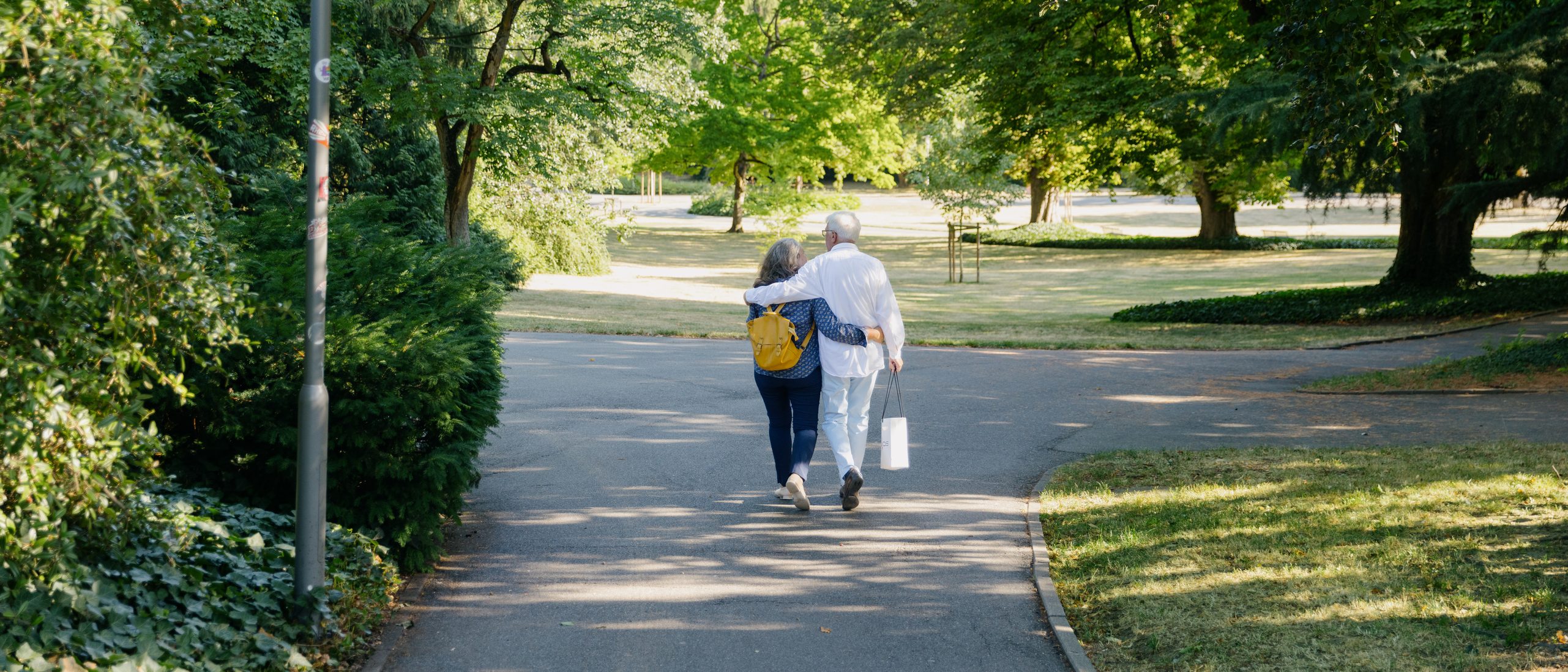 Eine Frau und ein Mann spazieren durch den Stadtpark, man sieht sie von hinten, rundherum Bäume, Wiese und eine Straßenlaterne.