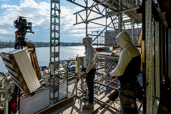 Deux ouvriers se tiennent en haut de l'échafaudage, à gauche la grue chargée, au fond le Rhin.