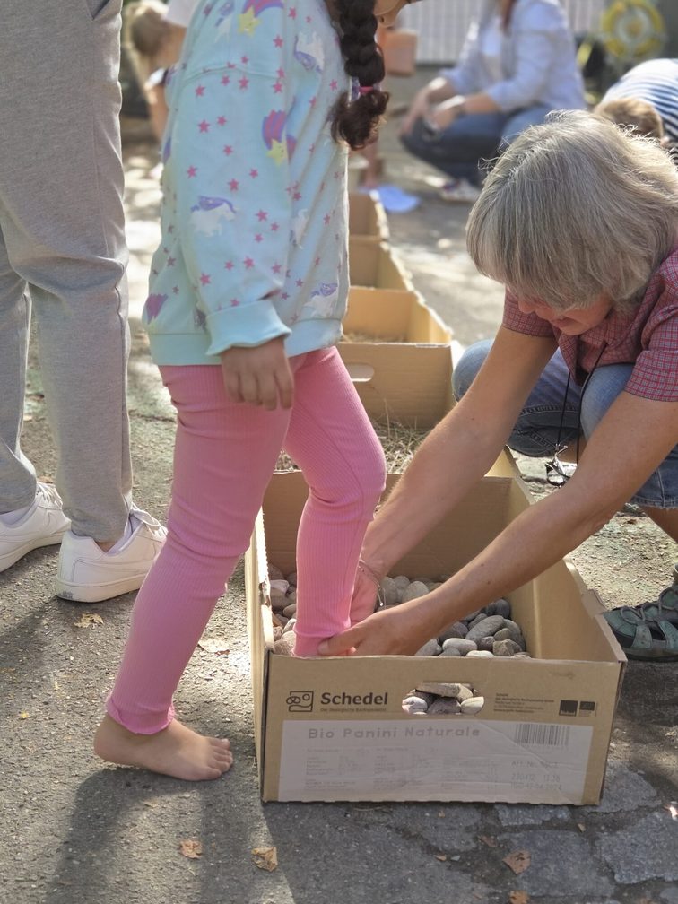 Kinderbeine, die mit der Hilfe von einer Erzieherin in eine Kiste eines Barfußpfades steigen