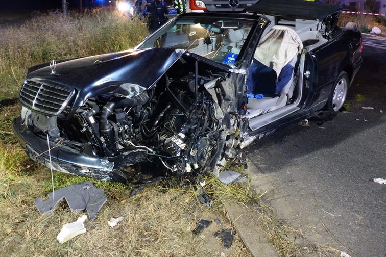 Demoliertes Unfallfahrzeug, Einsatzkräfte im Hintergrund.