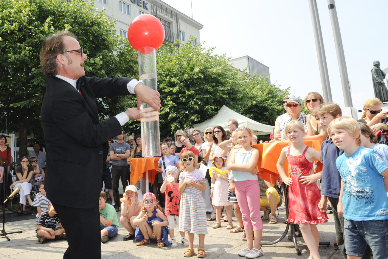 Wissenschaftsmarkt 2011 mit eoinem Mann im Vordergrund, der ein Experiment mit einer durchsichtigen Röhre durchführt, Kinder schauen zu.