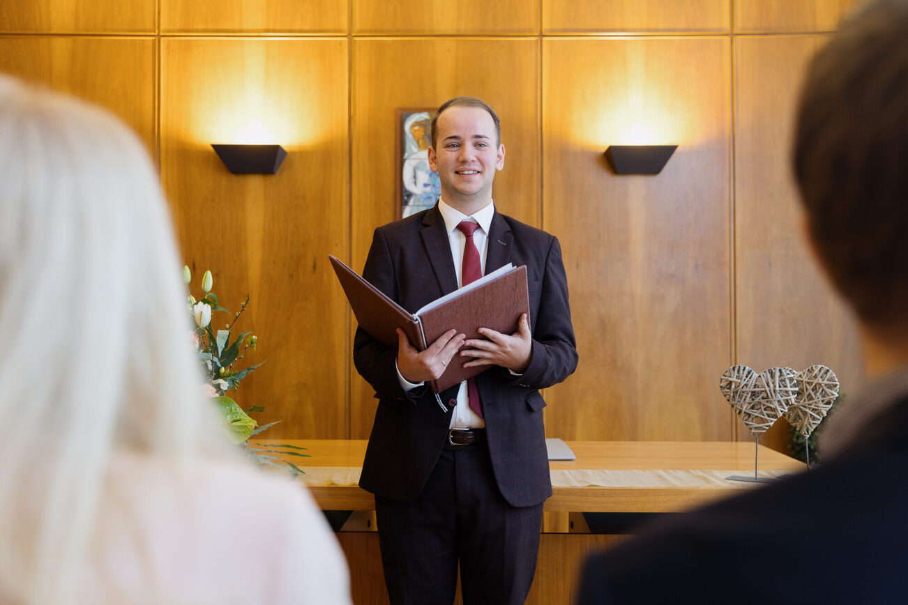 Ein Standesbeamter steht in der Mitte mit einem aufgeklappten braunen Buch, vorne links und rechts sind die Hinterköpfe einer Frau und eines Mannes zu erkennen.