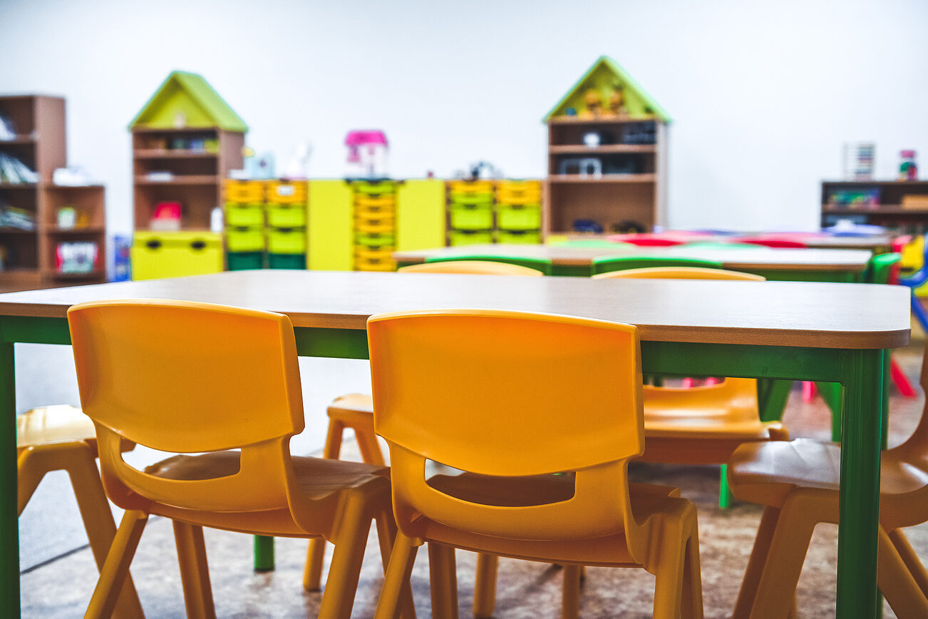 Chaises en plastique jaune autour d'une table au premier plan dans une crèche, avec en arrière-plan des étagères avec des casiers jaunes et verts.