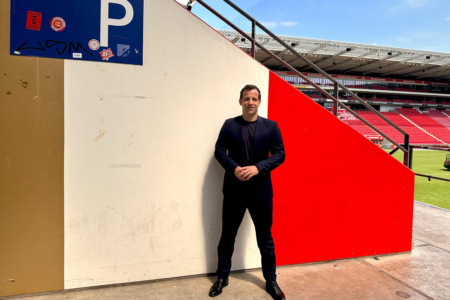 Oberbürgermeister Nino Haase steht vor dem P-Block in der Mewa Arena, seinem Lieblingsort in Mainz. Er schaut in die Kamera im Hintergrund ist ein kleines Stück des Spielfelds zu sehen, über der Arena ist blauer Himmel.