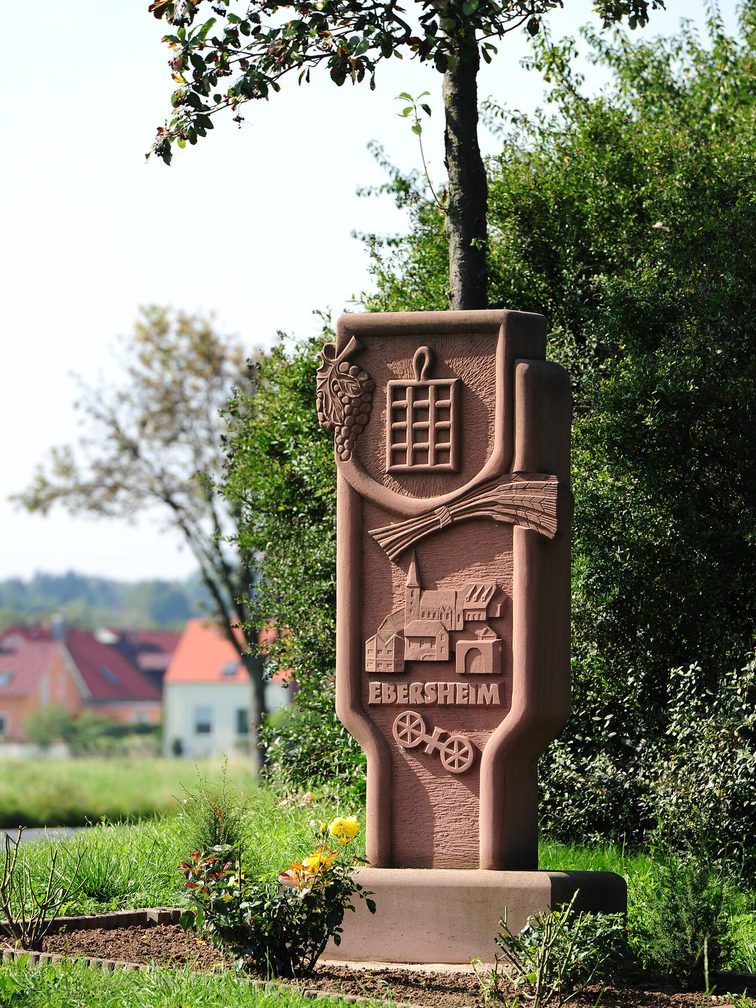 Monumento in pietra con l'iscrizione "Ebersheim", una piccola veduta della città, la ruota di Magonza e altri piccoli simboli